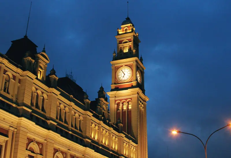 Fachada iluminada da Estação da Luz à noite, em São Paulo, com torre do relógio em destaque contra o céu azul escuro