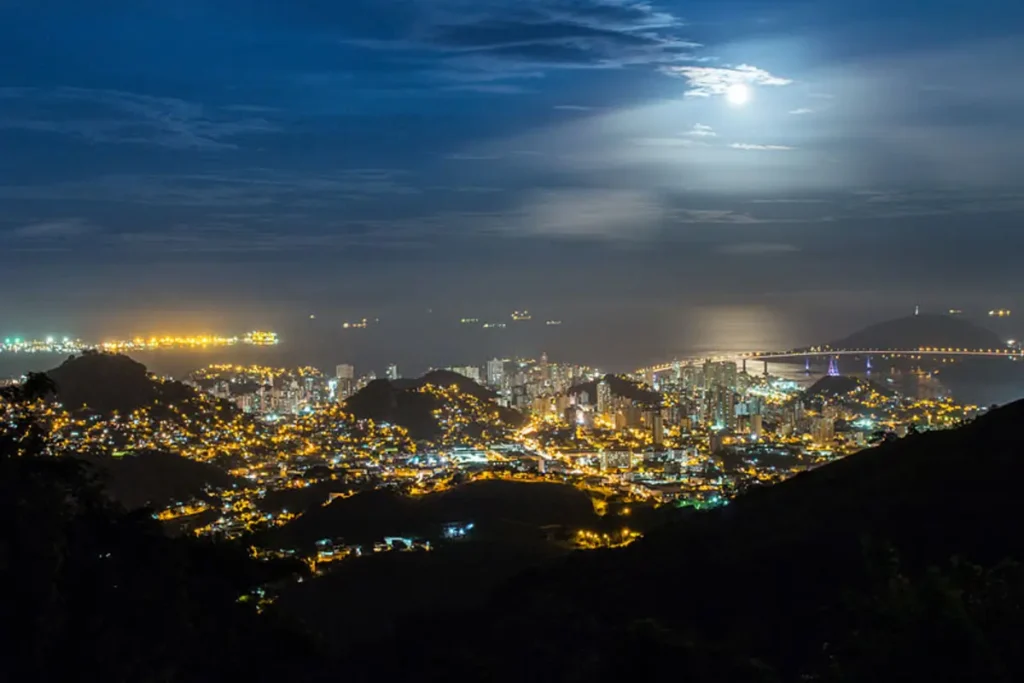 Vista noturna da Grande Vitória com a Terceira Ponte iluminada e a lua refletida no mar