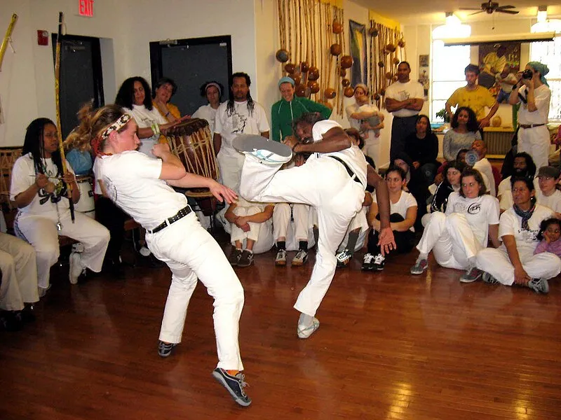 Roda de capoeira com mestres e alunos praticando movimentos em um salão de madeira