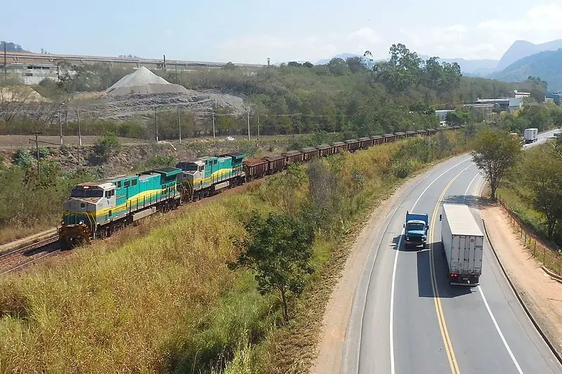 Trem de carga da Vale transportando minério paralelo a uma rodovia por onde passam caminhões, em região de mata e serras ao fundo