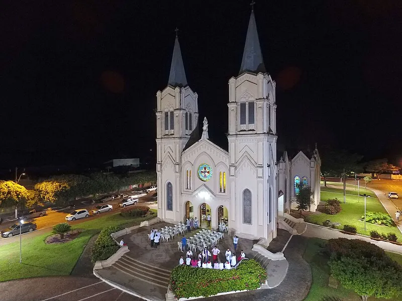 Fachada iluminada da Igreja Matriz de Aquidauana à noite, com fiéis reunidos em frente