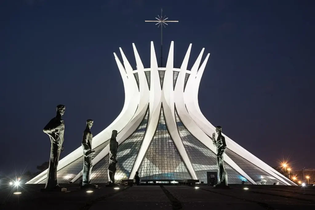 Fachada iluminada da Catedral Metropolitana de Brasília à noite, com esculturas na entrada. Por Rodrigo de Almeida Marfan, CC BY-SA 4.0, via Wikimedia Commons