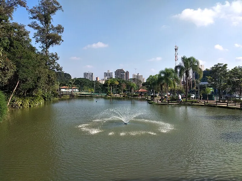 Lago com fonte no Bosque Maia em Guarulhos, cercado por árvores e prédios ao fundo