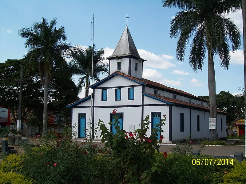 Igreja Matriz de Aparecida de Goiânia com fachada branca e detalhes em azul, rodeada por palmeiras e jardins floridos.