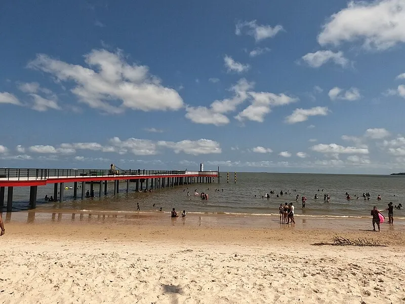 Praia com trapiche e banhistas em Barcarena, Pará, sob céu azul com nuvens
