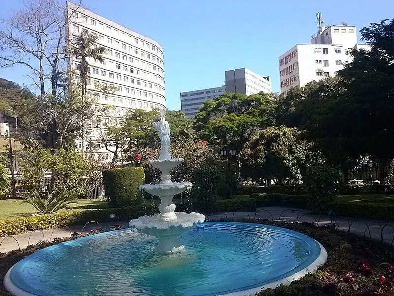 Fonte decorativa no centro de Petrópolis cercada por jardins e prédios ao fundo.